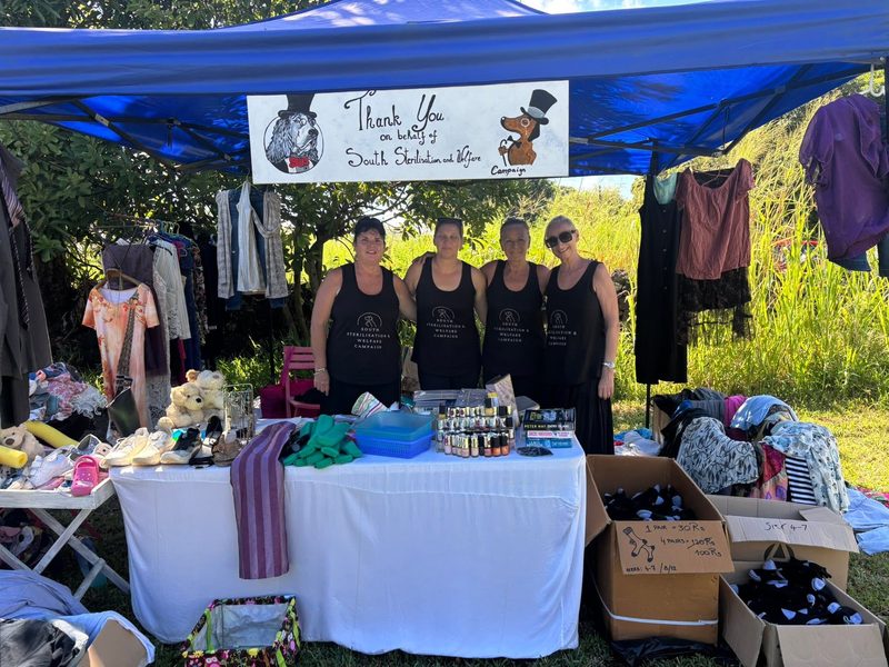 Four women in matching black tank tops stand at an outdoor charity stall with clothes and donated goods on tables
