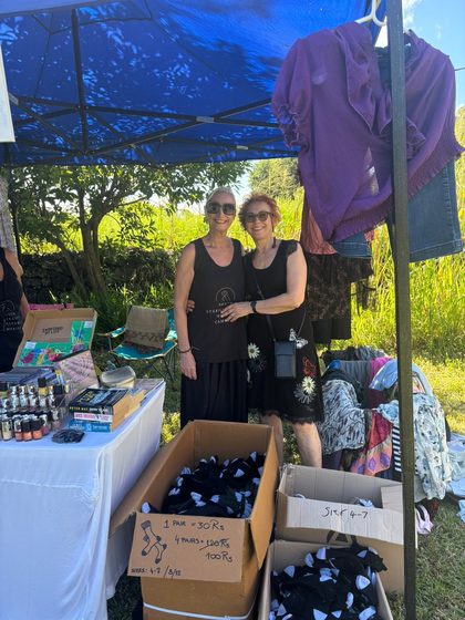 Two women stand smiling under a blue canopy at an outdoor market stall with clothes and goods for sale